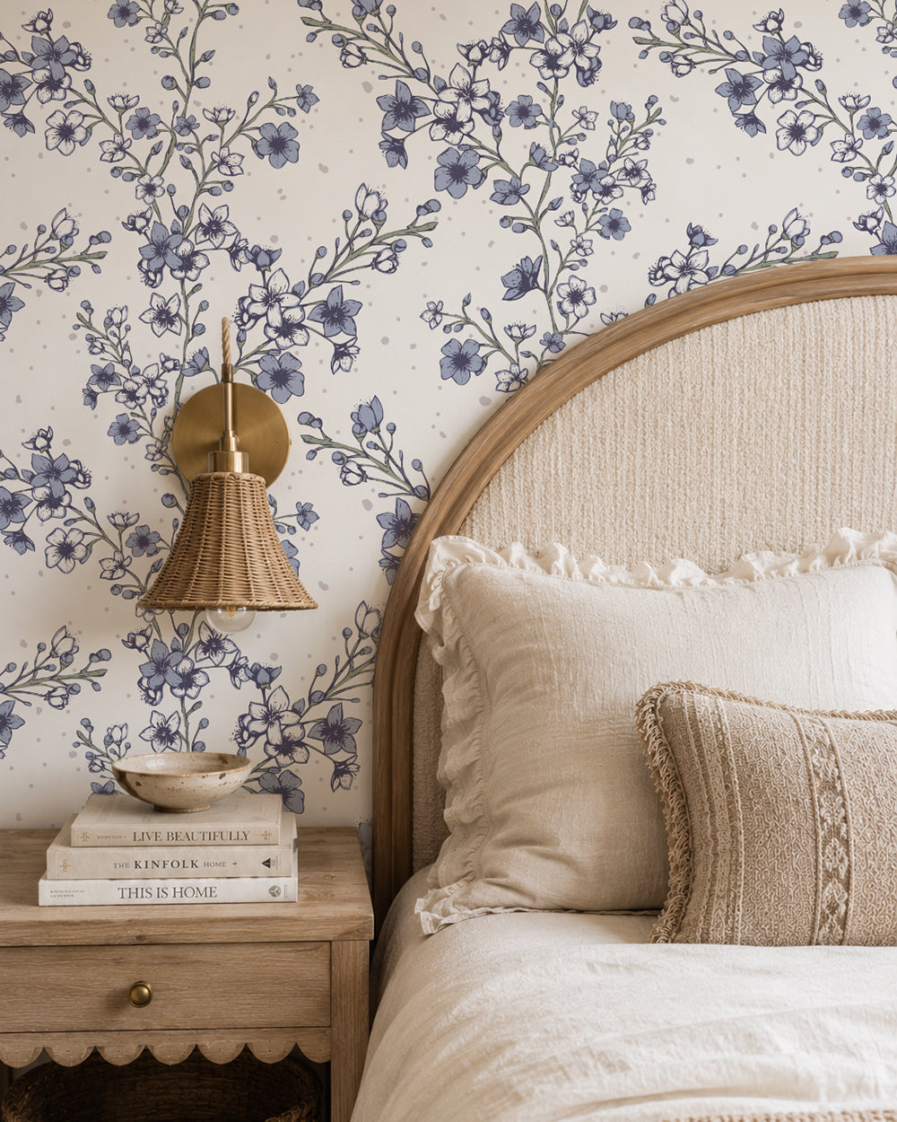 Blossom Whisper Dusty Blue floral wallpaper behind a curved wood headboard with cream bedding and a woven wall light.