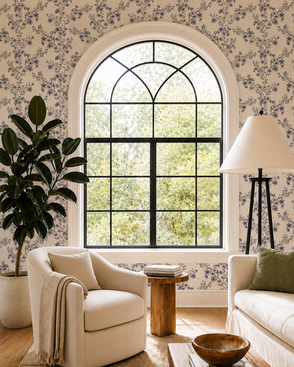 Blossom Whisper Dusty Blue floral wallpaper in a living room with an arched window, cream seating and natural wood accents.