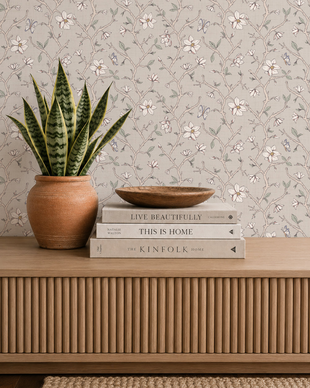 Magnolia Bloom Taupe wallpaper behind a wooden console styled with books, pottery and a potted plant.