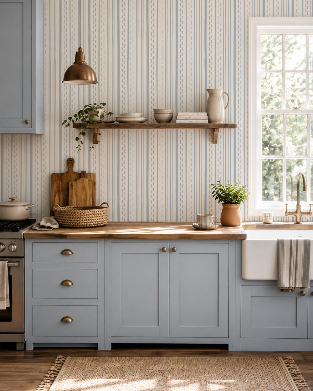 Ticking Stripe Dusty Blue on Cream wallpaper in a cozy kitchen with blue cabinets, wood counters and warm natural light.