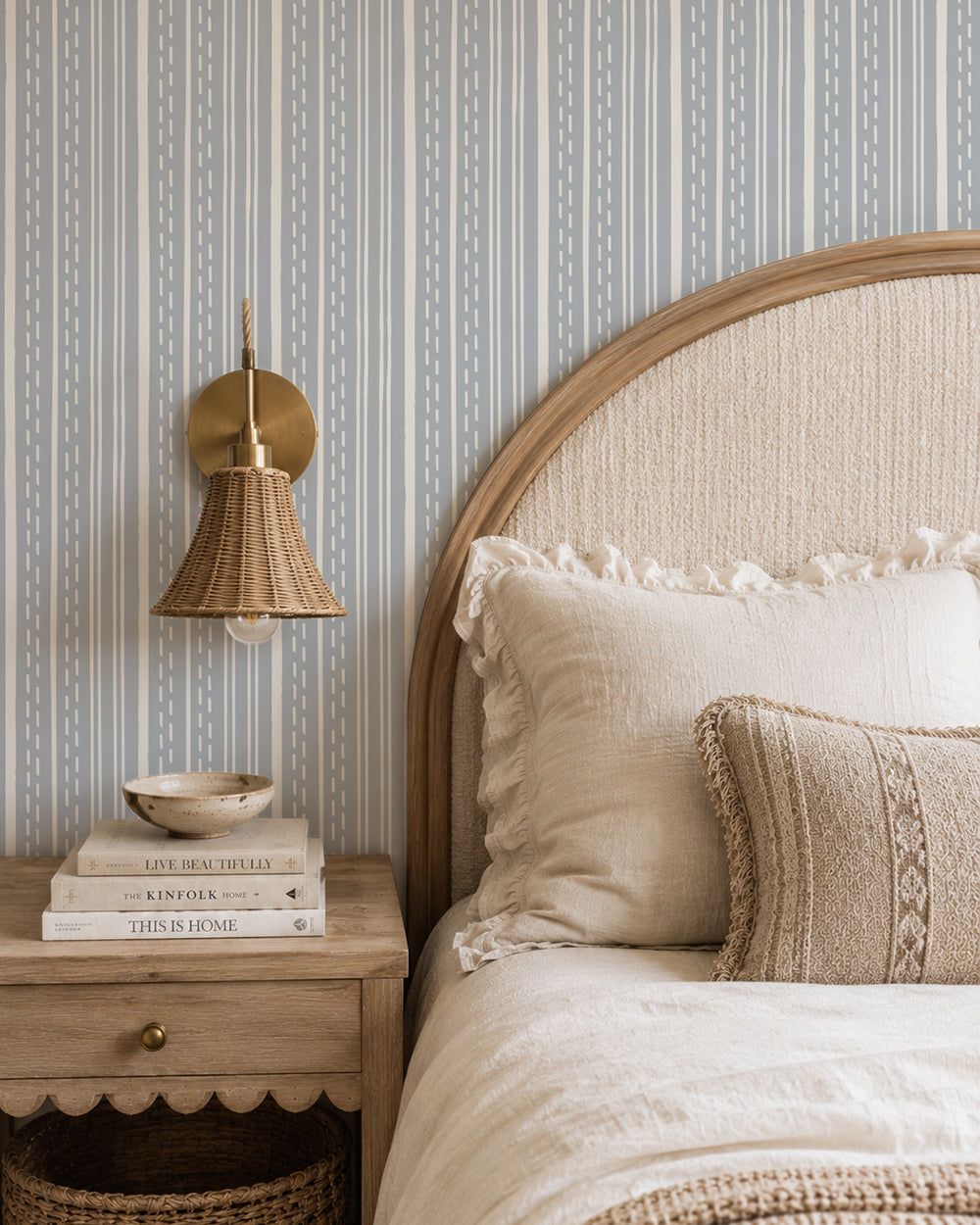 Ticking Stripe Dusty Blue wallpaper in a cozy bedroom with a curved wood headboard, cream bedding and woven lighting.